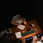 Man focused on work at an office desk, surrounded by gadgets and documents.